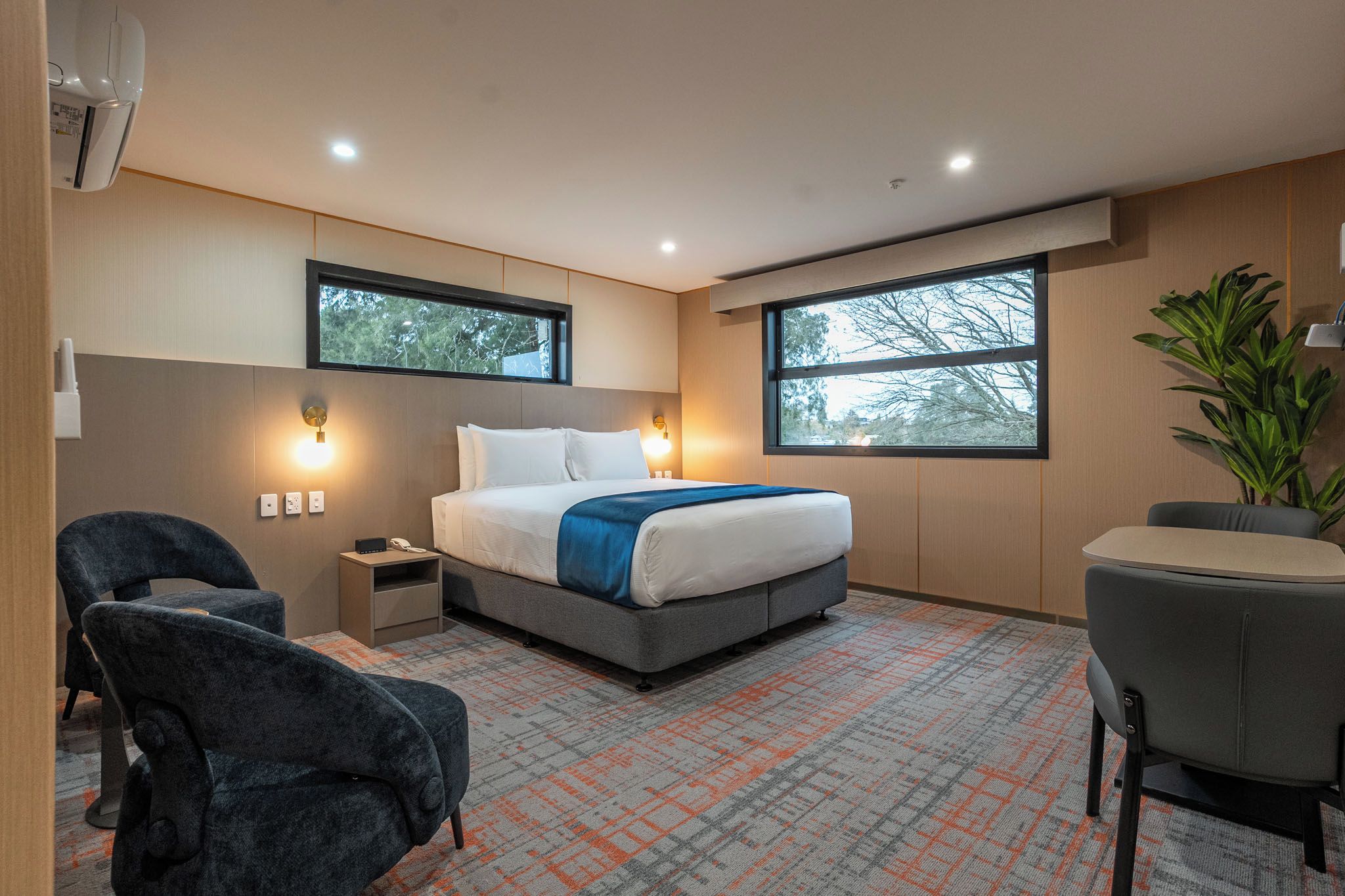 Habitación de hotel moderna con una cama grande, dos mesitas de noche, un escritorio y una silla. Hay dos ventanas que ofrecen luz natural y vistas a árboles.