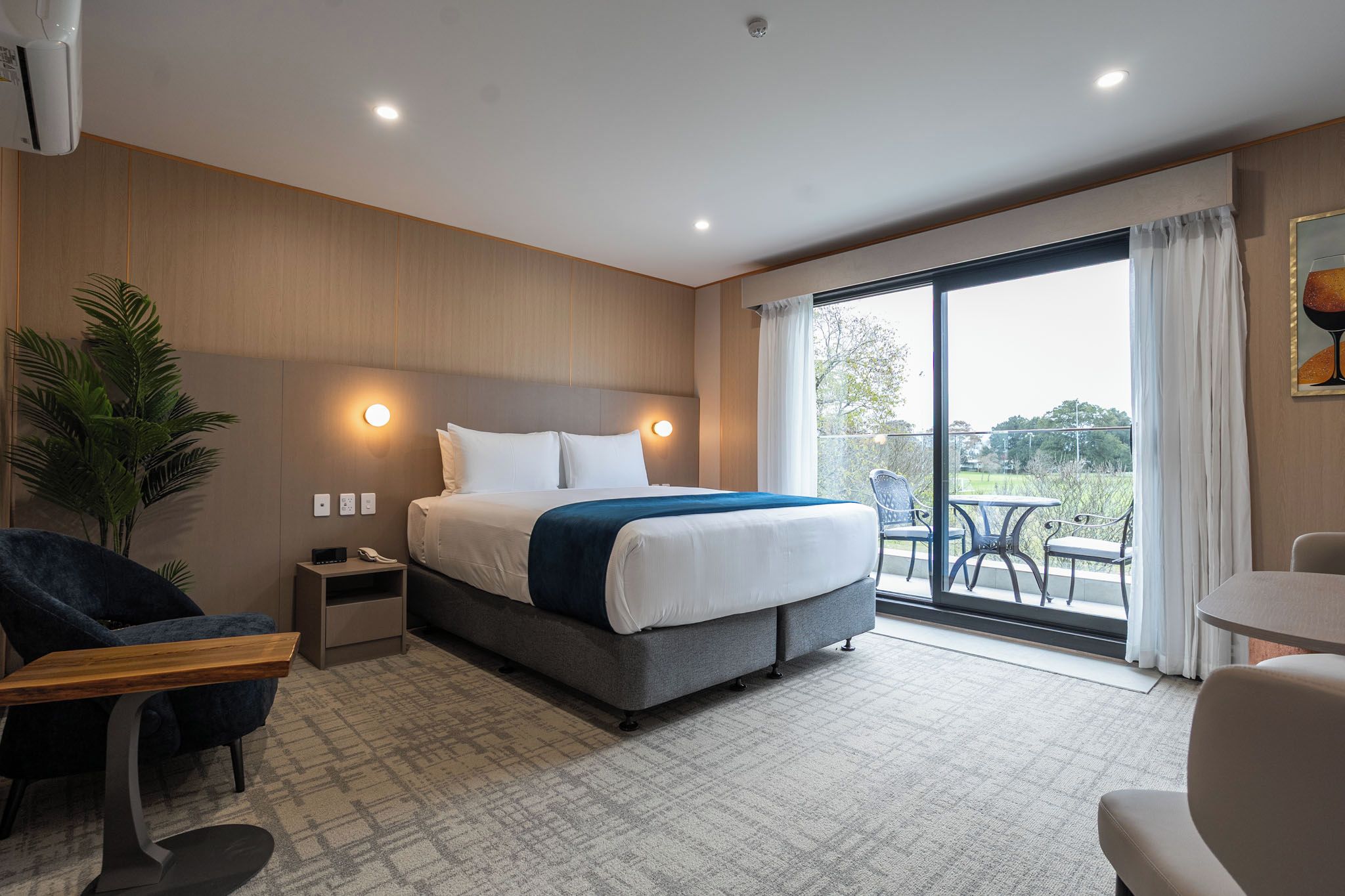 Habitación de hotel con cama, mesita de noche, lámpara, silla y vistas al exterior a través de un ventanal.