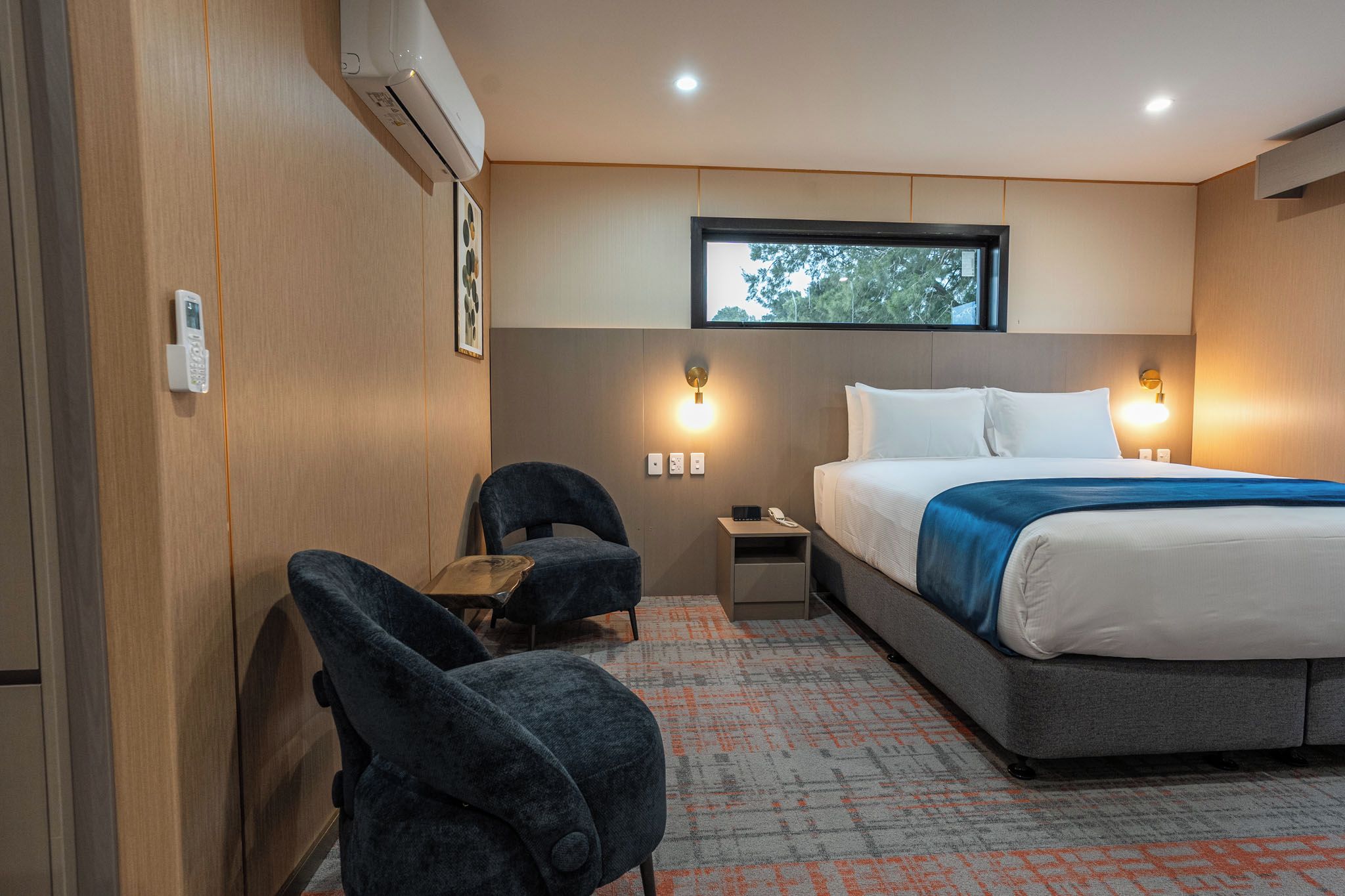 Habitació d'hotel amb un llit, dues cadires, un escriptori, una finestra i un aire condicionat fixat a la paret.