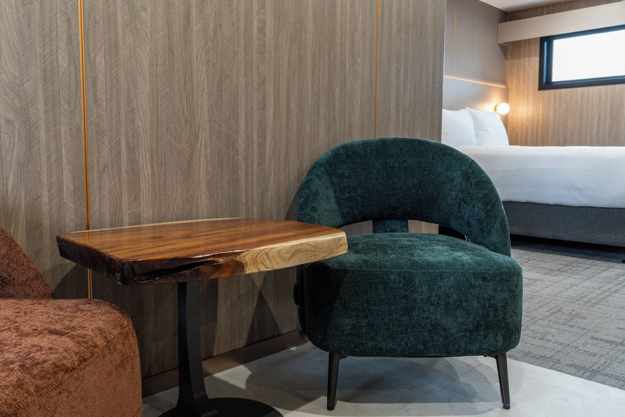 A hotel room with a wooden side table, a green upholstered chair, and a bed with white linens.