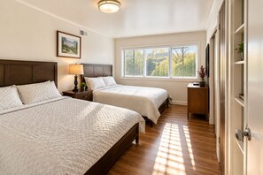 Un dormitorio con dos camas, una mesita de noche con lámpara, una ventana con persianas y una puerta adornada con una planta.