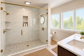 Un baño moderno con ducha envuelta en vidrio, paredes de mármol, un lavamanos blanco y una ventana con persianas.