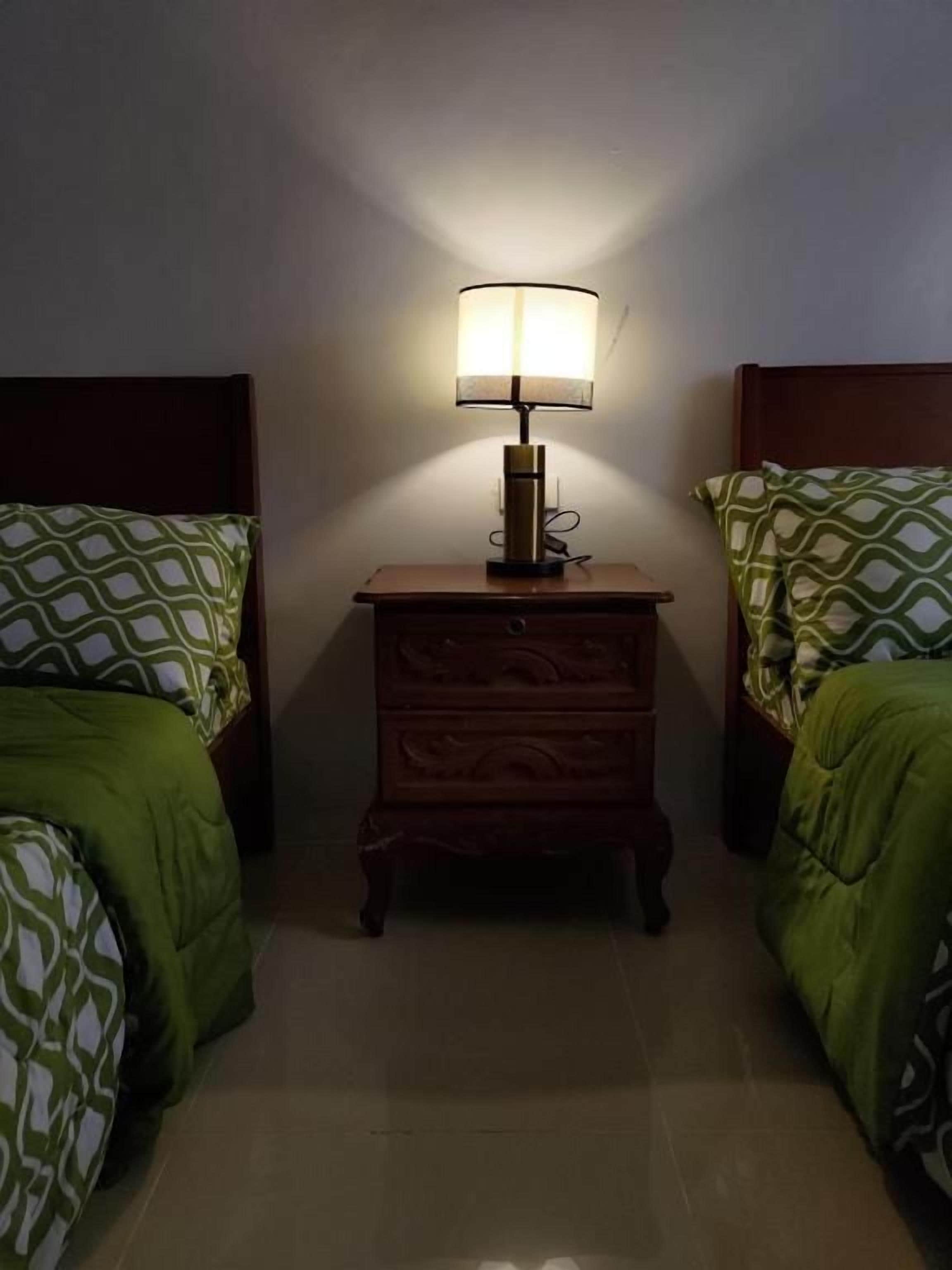 A room with two beds, each with patterned bedding, a nightstand with a lamp between them, and a wooden headboard.