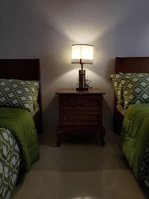 A room with two beds, each with patterned bedding, a nightstand with a lamp between them, and a wooden headboard. - JLF Inn (Daraga)
