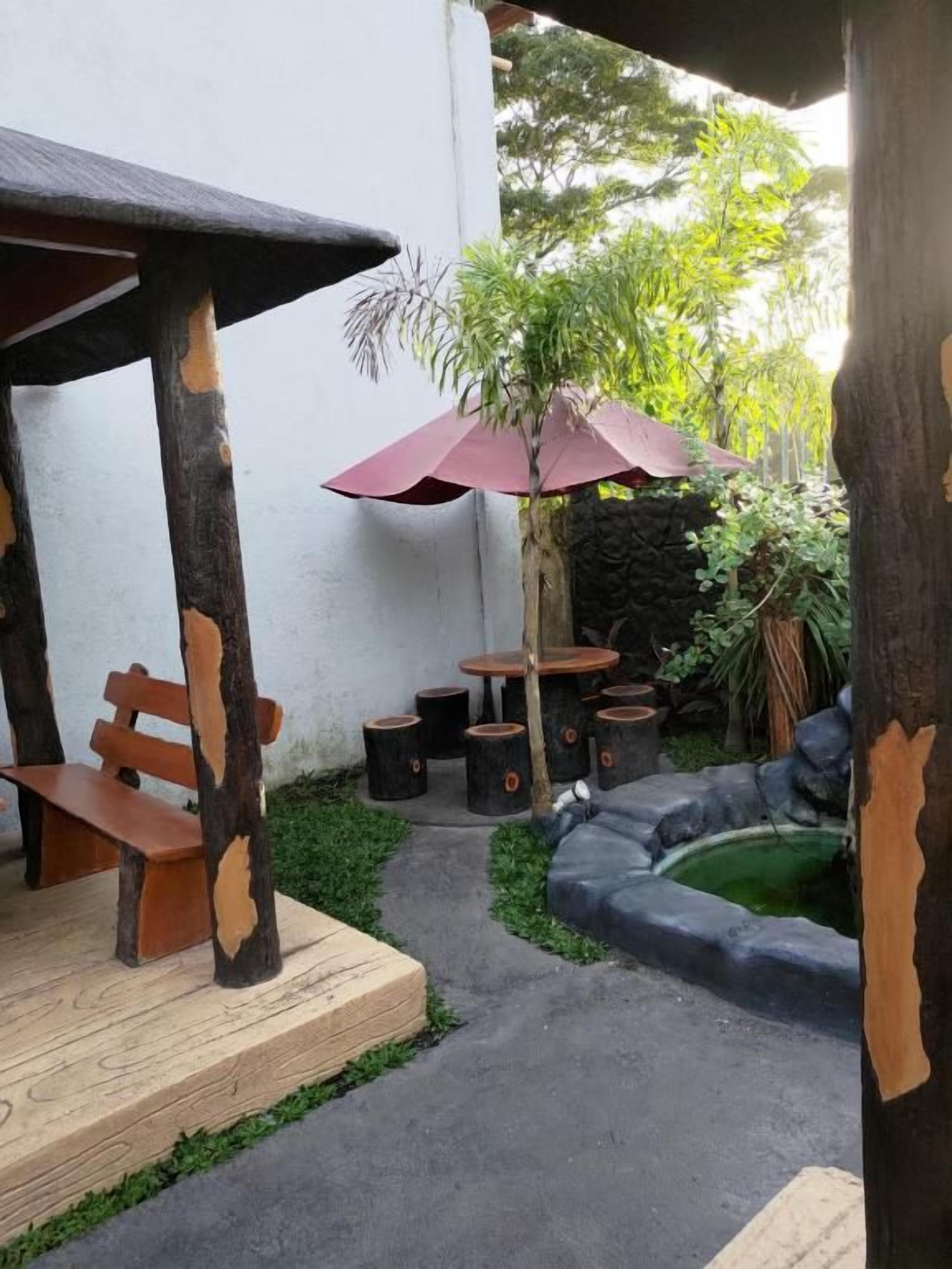 An outdoor seating area with wooden benches and tables, surrounded by greenery and a small pond.