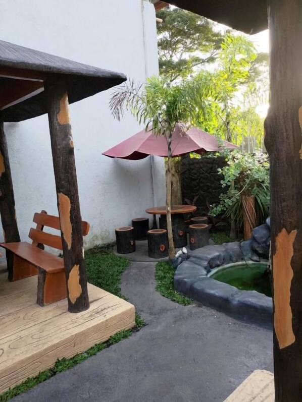 An outdoor seating area with wooden benches and tables, surrounded by greenery and a small pond.