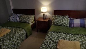 Habitación de hotel con dos camas, cada una con ropa de cama y almohadas estampadas. Entre las camas hay una mesita de noche con una lámpara.