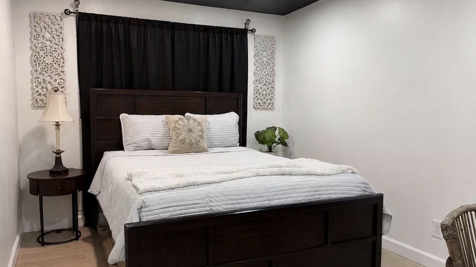 Ein Schlafzimmer mit einem dunkelholzbett, weißer Bettwäsche, einer Nachttischlampe, dekorativen Wandverkleidungen und einer Topfpflanze.
