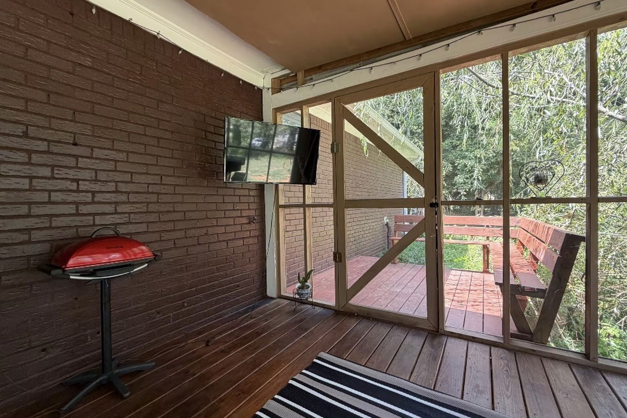 A room with a brick wall, a television mounted on the wall, a red barbecue grill, and a view of an outdoor deck with a wooden bench.