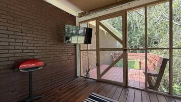A room with a brick wall, a television mounted on the wall, a red barbecue grill, and a view of an outdoor deck with a wooden bench.