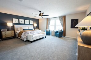 A bedroom with a bed, two side chairs, a nightstand, a lamp, a ceiling fan, and a window with curtains.