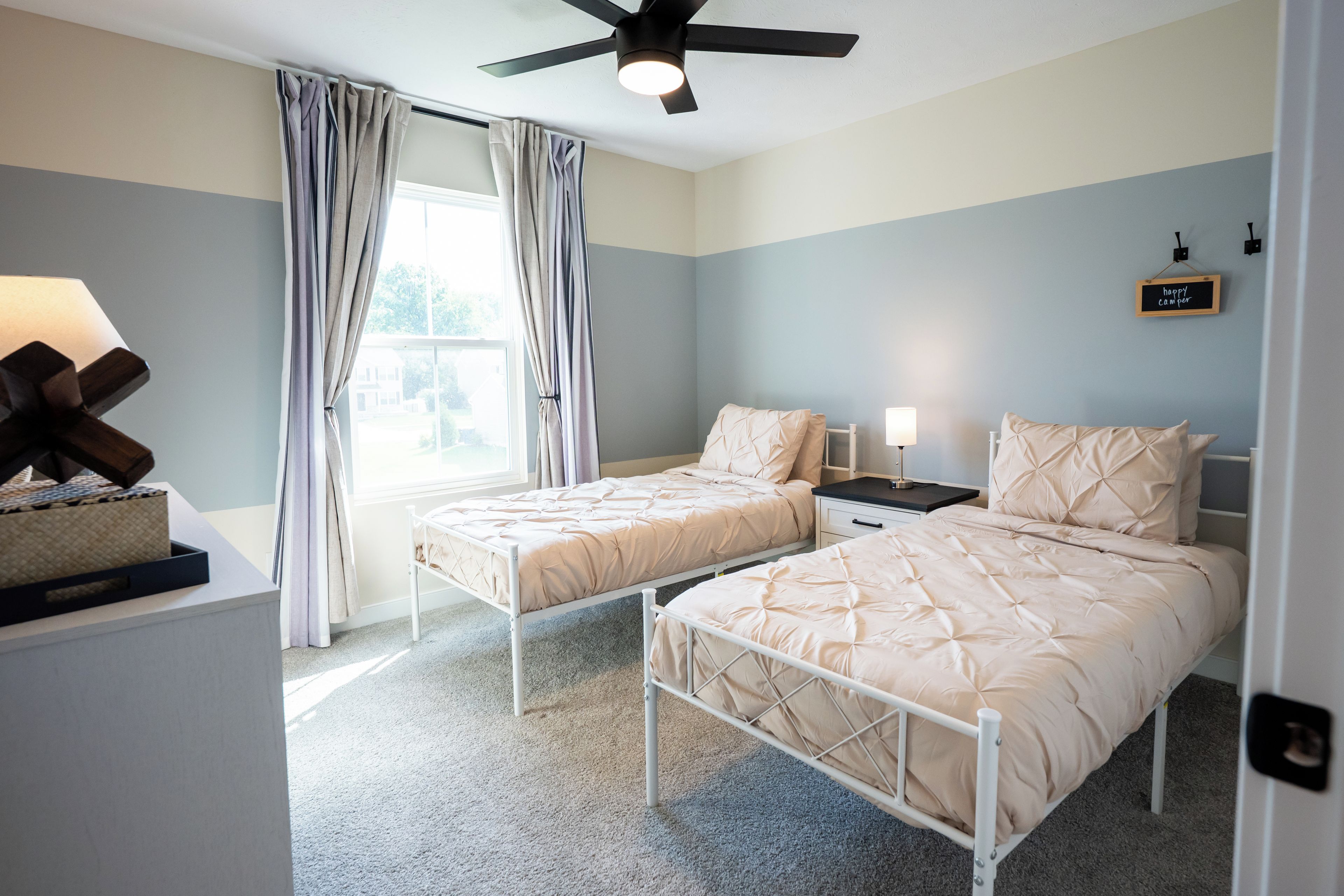 A bedroom with two beds, a ceiling fan, a bedside table with a lamp, and a window with curtains.