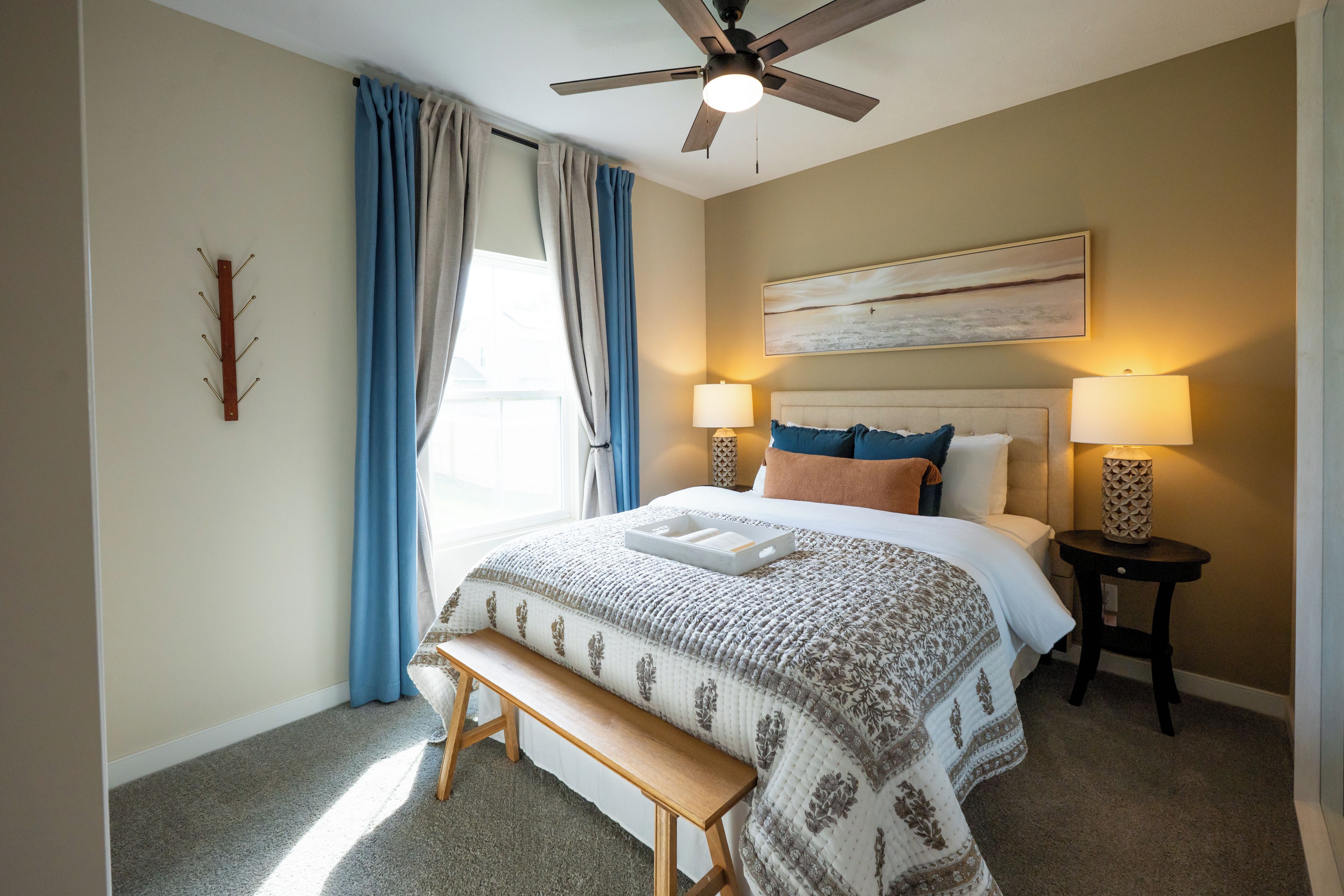 A bedroom with a bed, bedside lamps, a ceiling fan, a bench, and a painting on the wall.