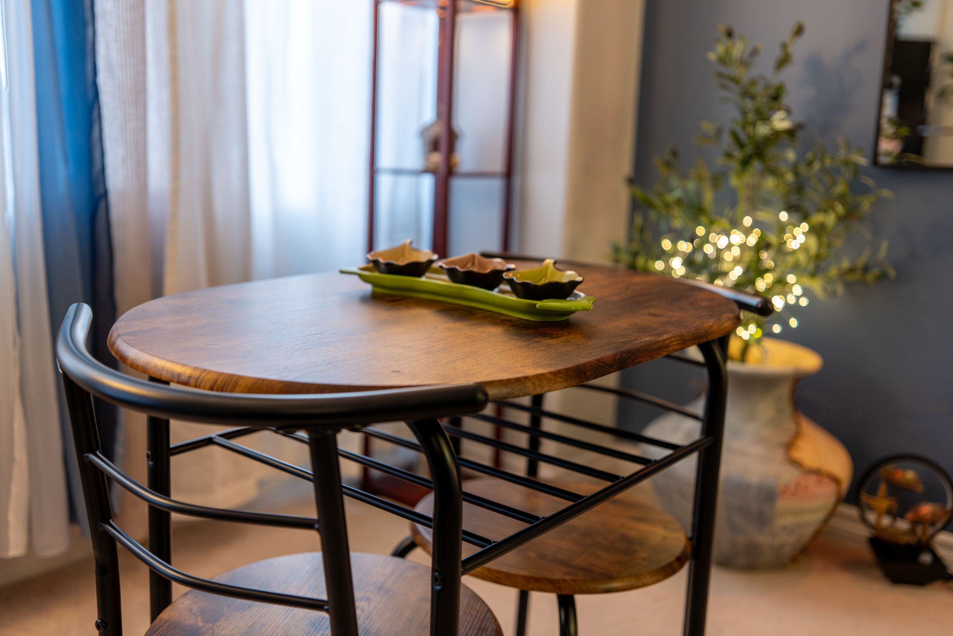A round wooden table with a black metal chair, a green plate with three bowls, and a potted plant with lights.