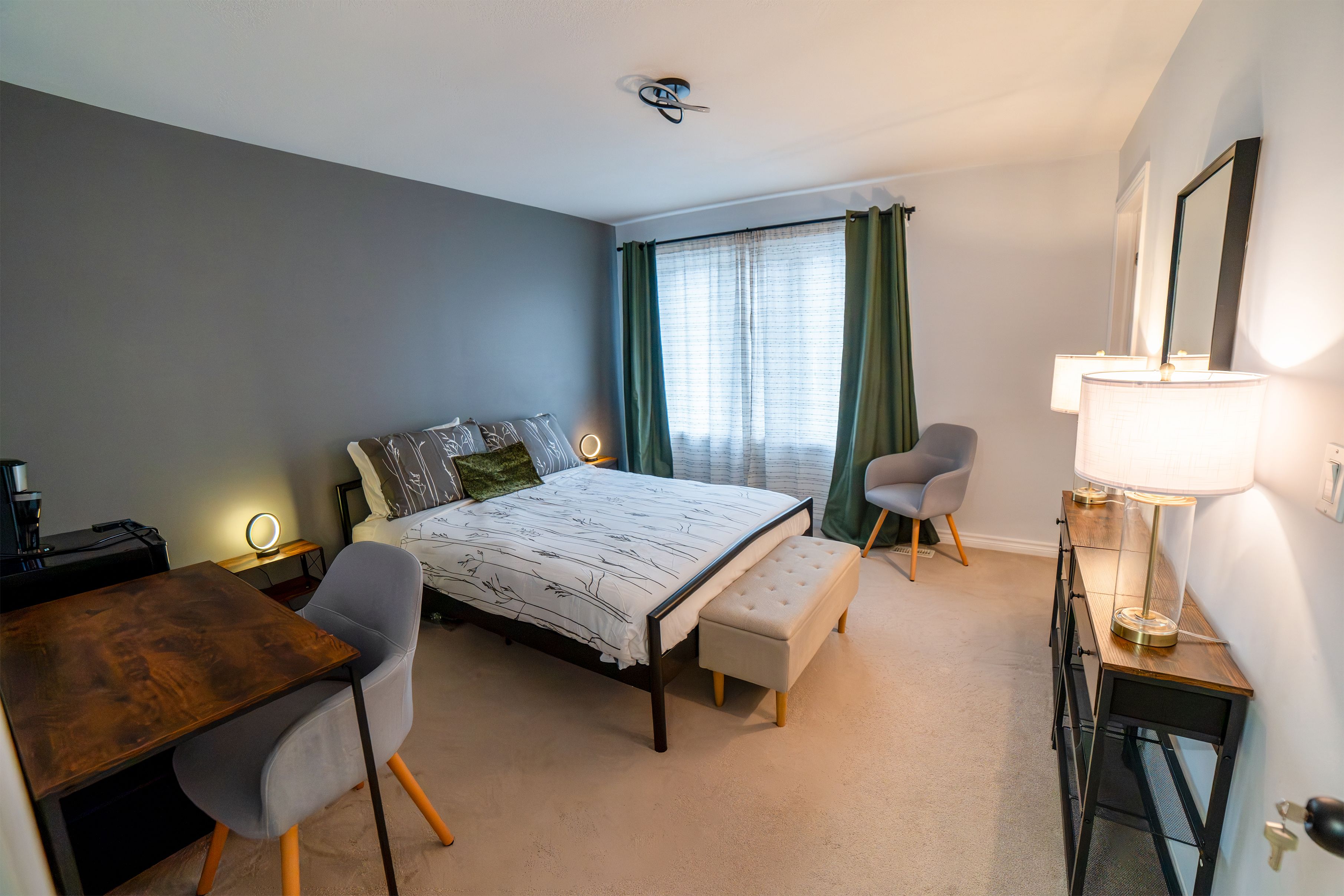 A hotel room with a bed, a desk, a chair, a bench, a dresser, and a mirror.