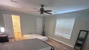 Un dormitorio con cama, ventilador de techo, ventana con persianas y puerta que da a otra habitación.