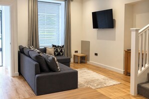 A modern living room with a grey sofa, a flat-screen TV mounted on the wall, and a wooden staircase.