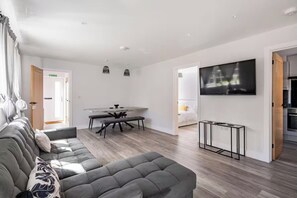 Ein modernes Wohnzimmer mit einem grauen Ecksofa, einem Esstisch mit Stühlen, einem an der Wand befestigten Flachbildfernseher und einer durch eine offene Tür einsehbaren Küchenzeile.