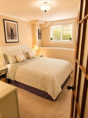 A bedroom with a large bed, a bedside table, a nightstand, a lamp, a framed picture on the wall, and a window with blinds. - The Maris l Lower Lonsdale Suite l Central & Cozy (North Vancouver)