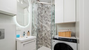 A modern bathroom with a glass shower enclosure, a built-in washing machine, and a vanity with a mirror. - Astronautów Chopin Airport Apartment (Warszawa)