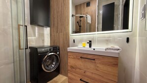 A modern bathroom with a glass shower enclosure, a washing machine, wooden cabinetry, and a large mirror. - Nyska Boutique Studio (Wrocław)