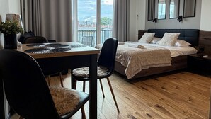 A modern bedroom with a wooden floor, a bed with a dark headboard, a dining table with chairs, and a view of a cityscape through the window. - Nyska Boutique Studio (Wrocław)