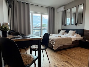Un dormitori modern amb sòl de fusta, un llit amb capçal de fusta fosc, una taula de menjador amb caires i una vista d'un paisatge urbà per la finestra.