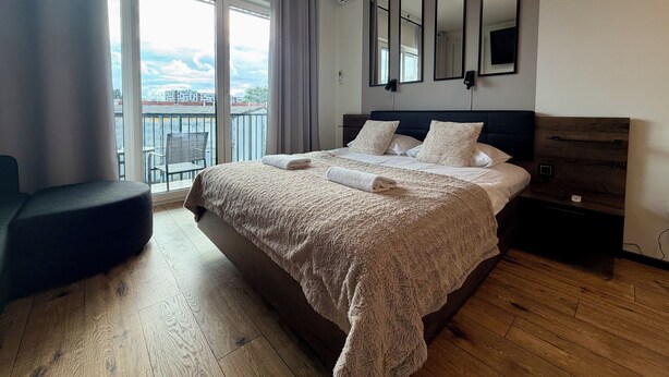 A modern hotel room with a large bed, a bedside table, a chair, and a view of the city through large windows. - Nyska Boutique Studio (Wrocław)