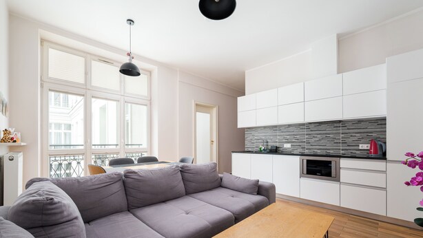Un soggiorno moderno con un divano grigio, un tavolino in legno e un angolo cottura con mobili bianchi e un forno a microonde.