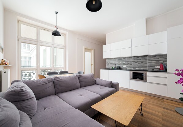 A modern living room with a grey sofa, a wooden coffee table, and a kitchen area with white cabinets and a microwave. - Metro Politechnika Spacious Apartment (Warszawa)
