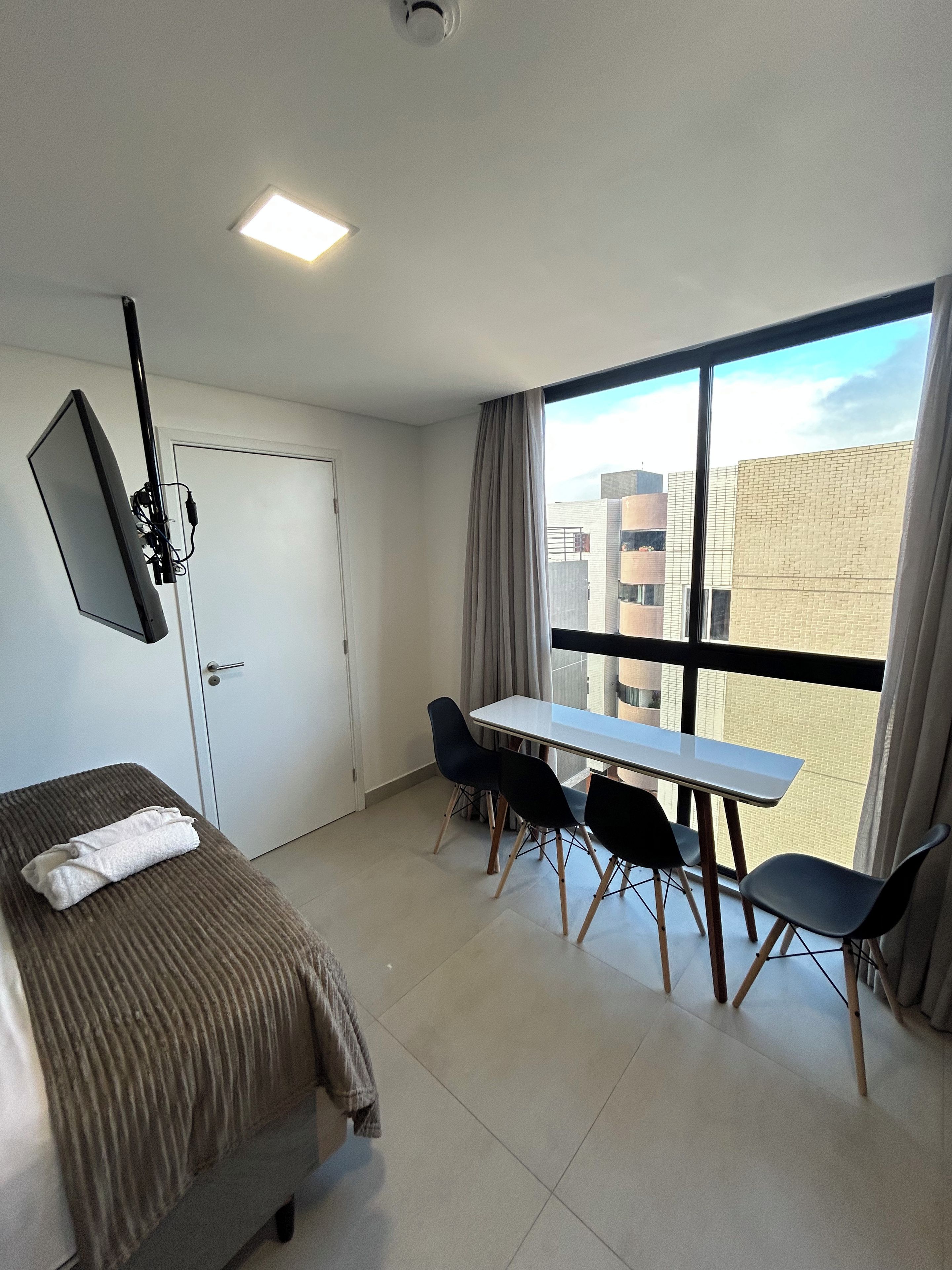 A hotel room with a bed, a desk with chairs, a large window, and a door.