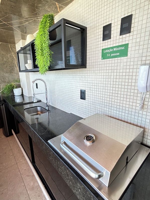 A modern kitchen with a stainless steel sink, a built-in grill, and a hanging plant. - Melhores Flats - Prático, 30m do mar (João Pessoa)