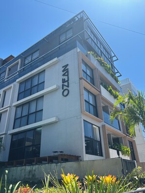 A multi-story building with a prominent 'NZOZEN' sign, large windows, and a balcony with greenery. - Melhores Flats - Prático, 30m do mar  (João Pessoa)