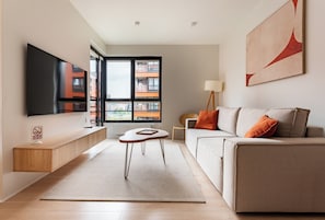 Uma sala de estar moderna com televisão de tela plana, um sofá com almofadas laranja, uma mesa de centro e uma janela grande com vista para edifícios.