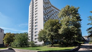 Un immeuble moderne de grande hauteur, doté d’une architecture originale aux lignes angulaires, entouré d’un parc bien entretenu, avec des arbres et une allée pavée.