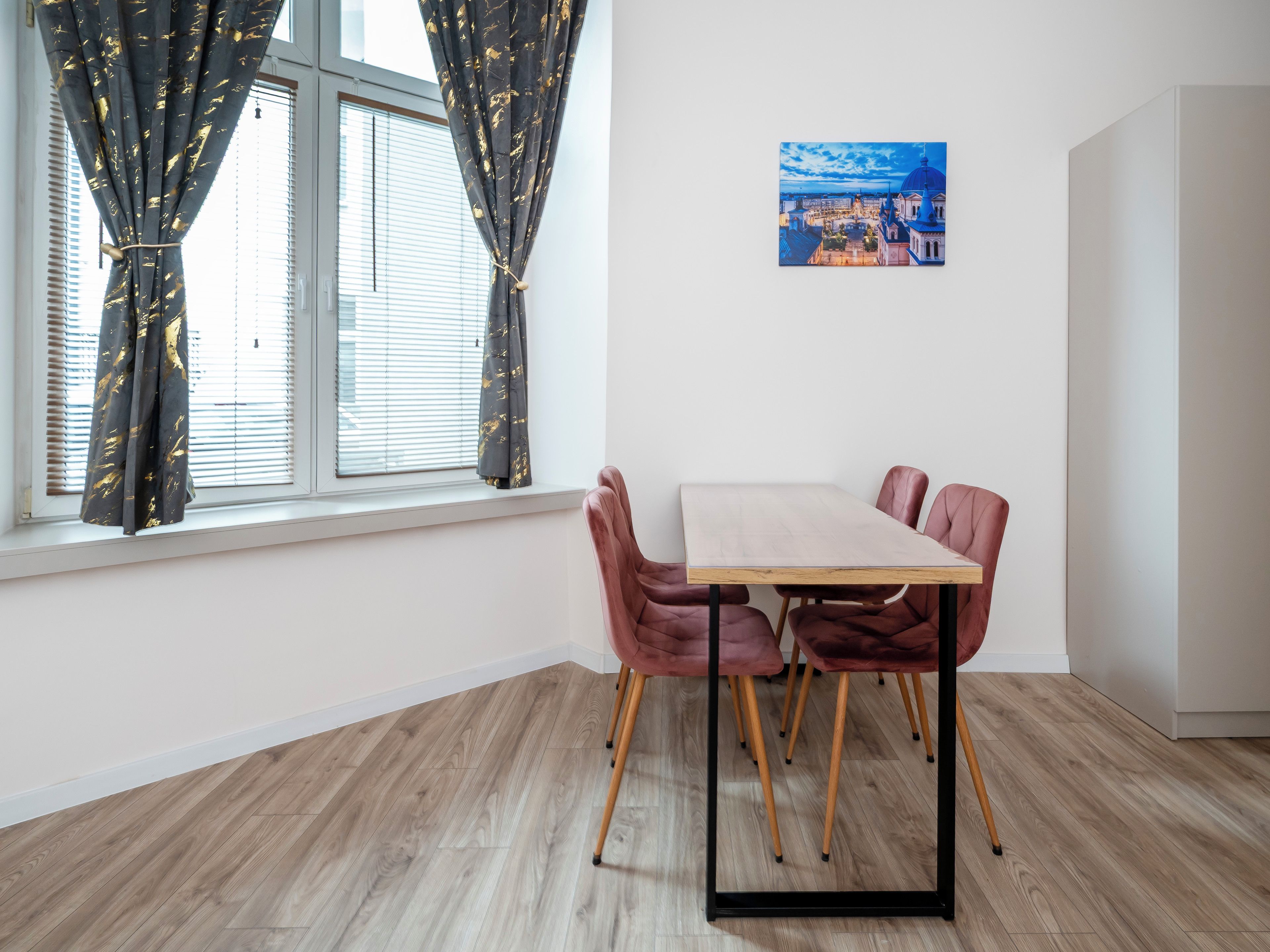 Une pièce comprenant une table à manger, des chaises, une fenêtre avec des rideaux et un tableau accroché au mur.