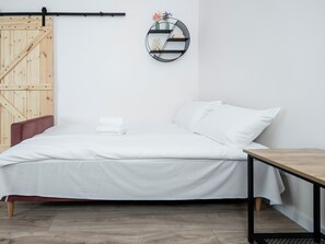 Un dormitori minimalista amb un llit, una porta de fusta, una prestatgeria rodona i una taula petita.