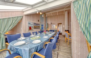 A dining area with a long table set for a meal, blue upholstered chairs, and wooden panel walls. - Nice home in Otterndorf with sauna (Otterndorf)