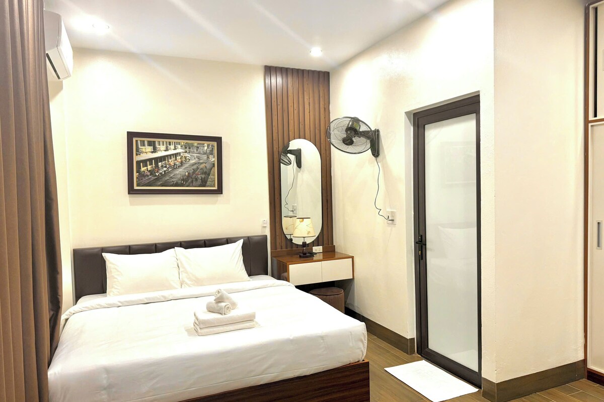 A hotel room with a bed, a bedside table with a lamp, a wall-mounted fan, a framed picture, and a sliding door.