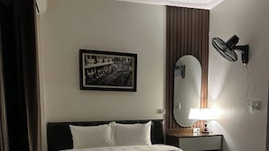 Ein Hotelzimmer mit einem Bett, einem Wandspiegel, einem Bild, einem Standventilator und einem Nachttisch mit Lampe.