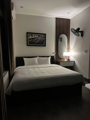 A hotel room with a bed, a wall-mounted mirror, a picture, a standing fan, and a bedside table with a lamp. - NESTA HOTEL  (Hoa Lu)