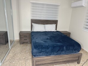 Ein Schlafzimmer mit einem Bett, einem Nachttisch, einem Spiegel und einem Fenster mit Jalousien.