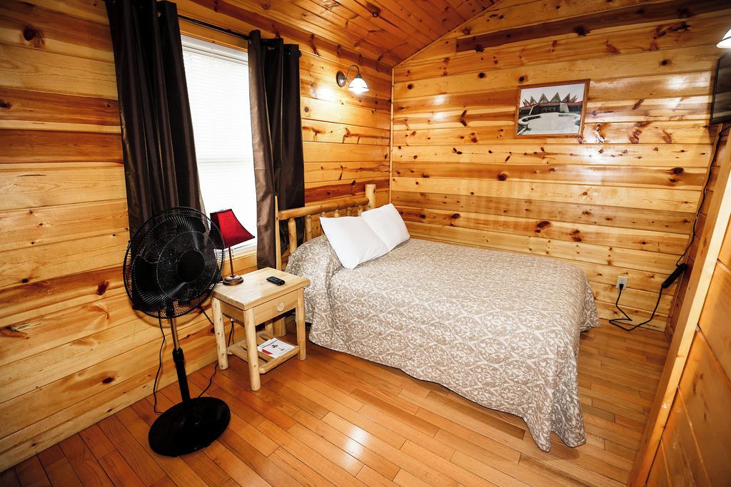 A wooden-paneled room with a bed, a nightstand, a fan, and a window with curtains.