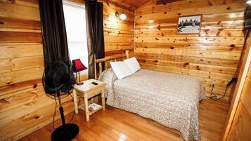 A wooden-paneled room with a bed, a nightstand, a fan, and a window with curtains.
