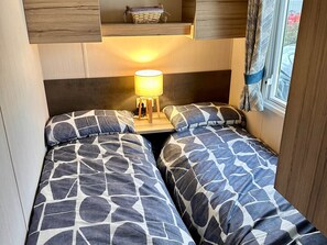 A small, cozy room with two beds, a nightstand with a lamp, and a window with curtains. - Cabin Sleeps 6 with Dishwasher (Skegness)