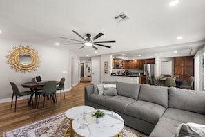 A modern living room with a grey sofa, a dining table with chairs, a ceiling fan, and a kitchen area with wooden cabinets and a refrigerator. - Pool & Spa • Game Room • King Beds • 20 Guests Max (Las Vegas)