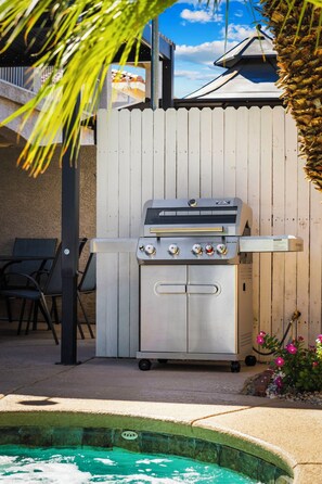 A portable stainless steel grill with four control knobs, positioned in front of a wooden fence with a pool in the background. - Pool & Spa • Game Room • King Beds • 20 Guests Max (Las Vegas)