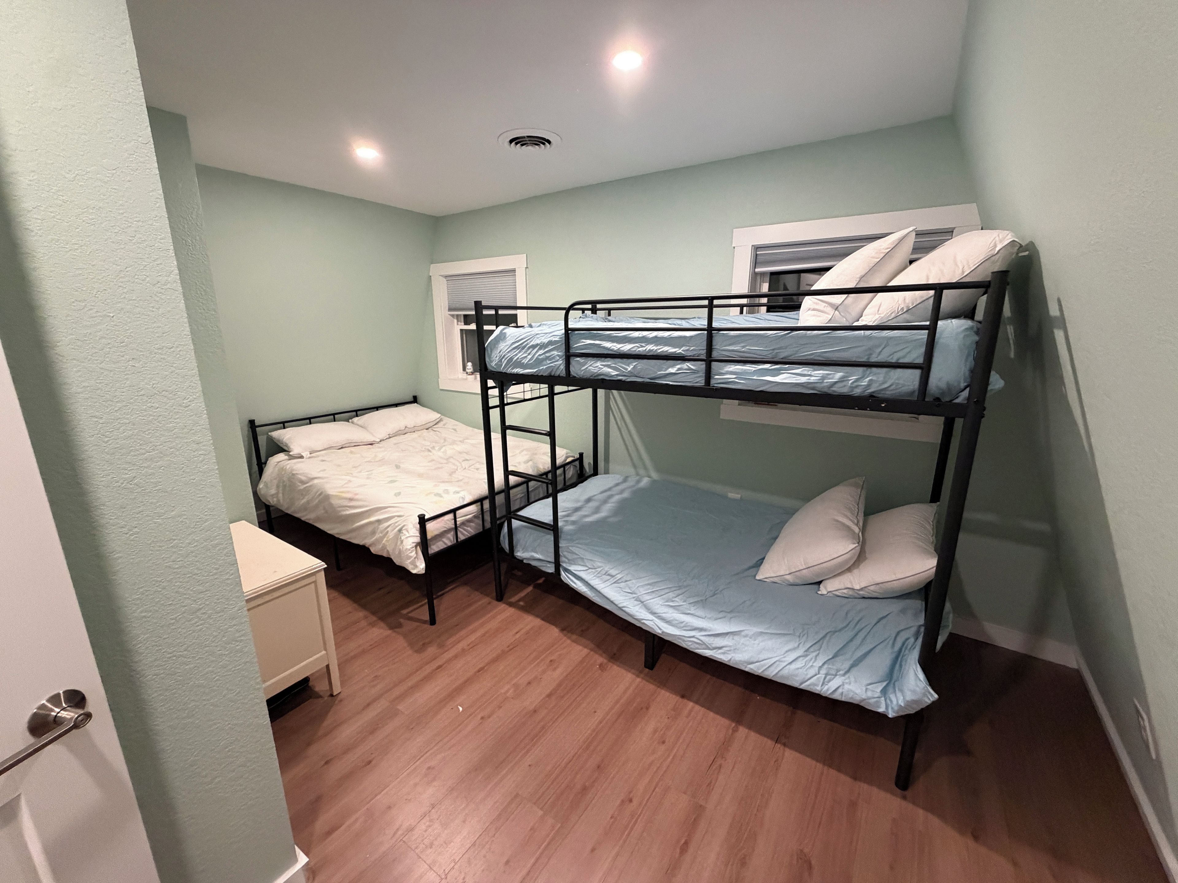 A bunk bed room with a wooden floor, a small bedside table, and a window with blinds.