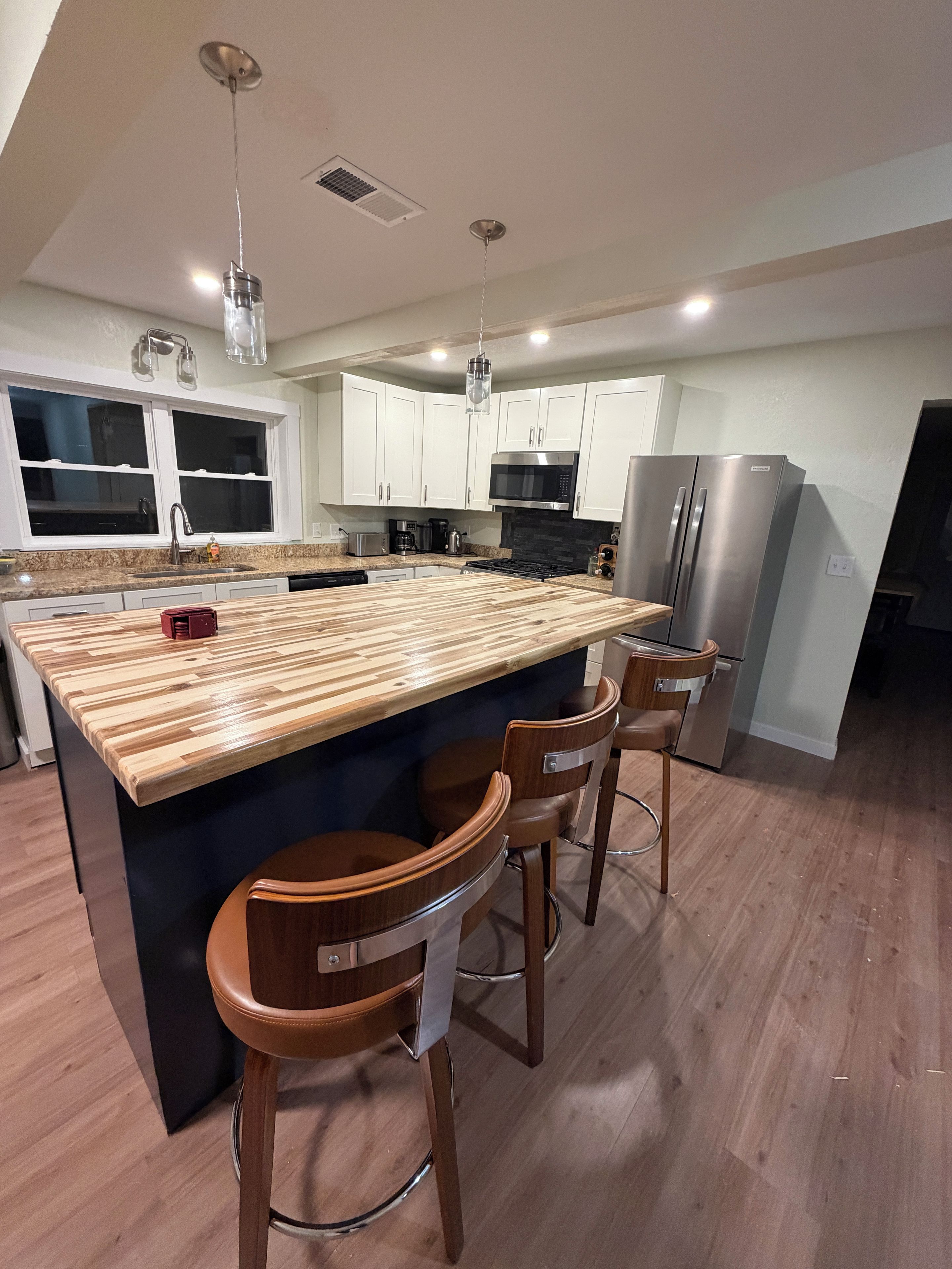 A modern kitchen with a central island, bar stools, stainless steel appliances, and a window overlooking the room.