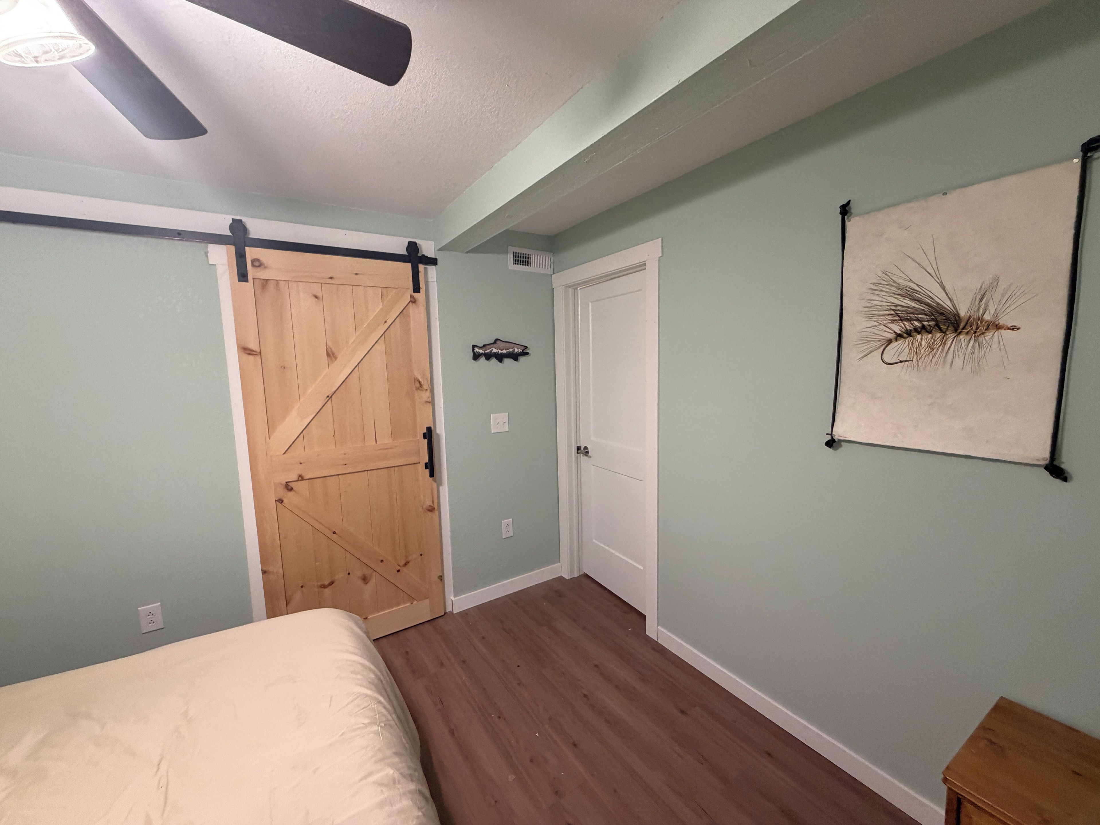 A bedroom with a bed, a wooden door, a white door, a ceiling fan, and a wall-mounted decoration.